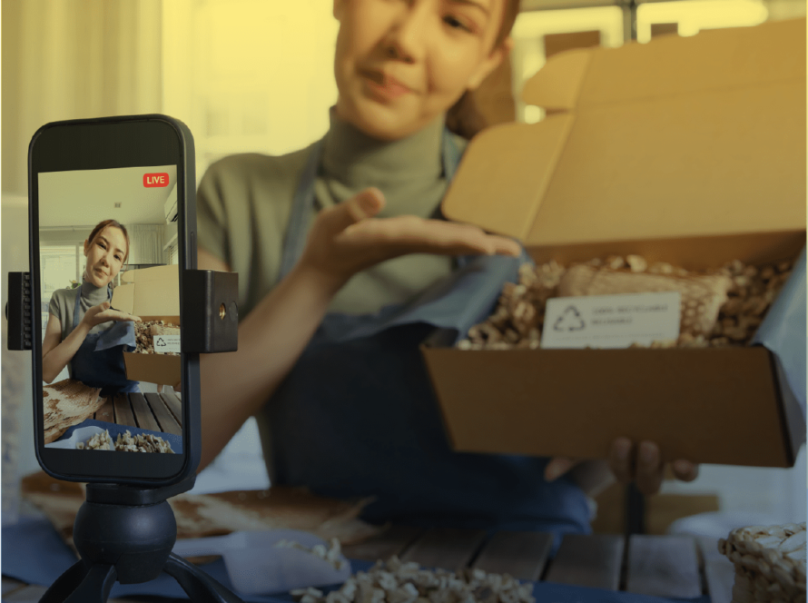 Eine Frau präsentiert ein geöffnetes Paket während eines Livestreams. Sie zeigt den Inhalt der Box, der mit recycelbaren Materialien verpackt ist. Ein Smartphone auf einem Stativ filmt die Szene. Sie ist gerade dabei User Generated Content zu produzieren.